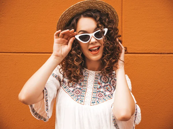 Beautiful smiling model with afro curls hairstyle dressed in summer hipster white dress and sunglasses.Carefree girl posing in the street near orange wall in hat.Funny and positive woman having fun