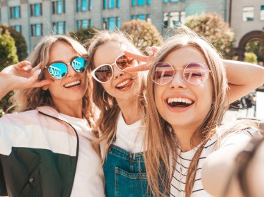 Yazlık kıyafetler giyen üç genç gülümseyen hipster kadın. Akıllı telefonda selfie selfie portre fotoğrafları çeken kızlar. Sokakta poz veren mankenler. Kadın güneş gözlüğü olumlu yüz duyguları gösteren