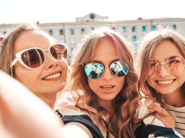Yazlık kıyafetler giyen üç genç gülümseyen hipster kadın. Akıllı telefonda selfie selfie portre fotoğrafları çeken kızlar. Sokakta poz veren mankenler. Kadın güneş gözlüğü olumlu yüz duyguları gösteren
