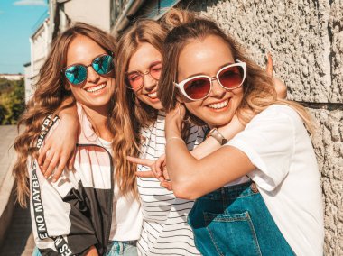 Moda yazlık giysiler üç genç güzel gülümseyen hipster kız Portresi. Seksi kaygısız kadınlar duvara yakın sokakta poz. Güneş gözlüğü ile eğlenen pozitif modeller. Sarılma