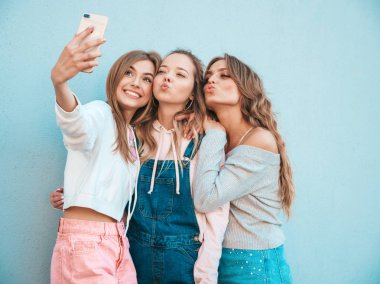 Yazlık kıyafetler giyen üç genç gülümseyen hipster kadın. Akıllı telefonda selfie selfie portre fotoğrafları çeken kızlar. Duvara yakın sokakta poz veren mankenler. Kadın olumlu yüz duyguları gösteren