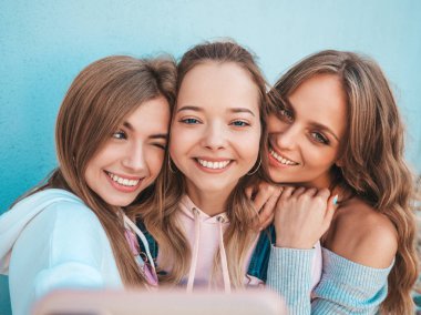 Yazlık kıyafetler giyen üç genç gülümseyen hipster kadın. Akıllı telefonda selfie selfie portre fotoğrafları çeken kızlar. Duvara yakın sokakta poz veren mankenler. Kadın olumlu yüz duyguları gösteren