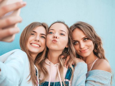 Yazlık kıyafetler giyen üç genç gülümseyen hipster kadın. Akıllı telefonda selfie selfie portre fotoğrafları çeken kızlar. Duvara yakın sokakta poz veren mankenler. Kadın olumlu yüz duyguları gösteren