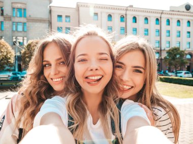 Yazlık kıyafetler giyen üç genç gülümseyen hipster kadın. Akıllı telefonda selfie selfie portre fotoğrafları çeken kızlar. Sokakta poz veren mankenler. Kadın olumlu yüz duyguları gösteren 