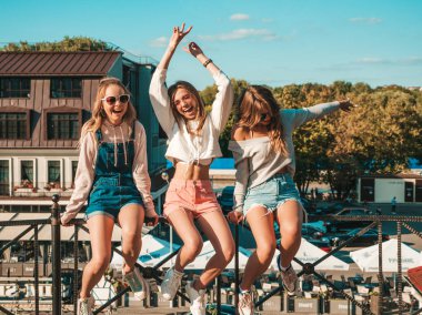Moda yazlık giysiler üç genç güzel gülümseyen hipster kız Portresi. Sokakta ki tırabzanda oturan seksi kaygısız kadınlar. Güneş gözlüğü ile eğlenen pozitif modeller. El kaldırıyorlar.