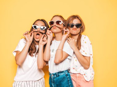 Moda yazlık giysiler üç genç güzel gülümseyen hipster kız. Seksi kaygısız kadınlar stüdyoda sarı duvar yakınında poz. Pozitif modeller deli gidiyor ve eğleniyor. Saç ile bıyık yapma