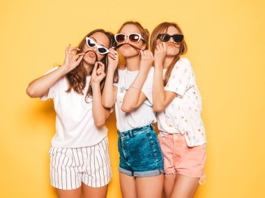 Moda yazlık giysiler üç genç güzel gülümseyen hipster kız. Seksi kaygısız kadınlar stüdyoda sarı duvar yakınında poz. Pozitif modeller deli gidiyor ve eğleniyor. Saç ile bıyık yapma