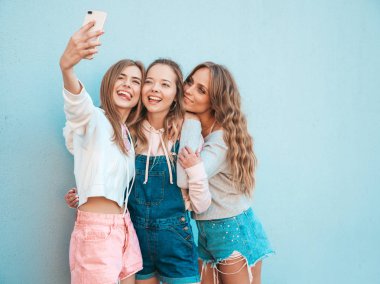 Yazlık kıyafetler giyen üç genç gülümseyen hipster kadın. Akıllı telefonda selfie selfie portre fotoğrafları çeken kızlar. Duvara yakın sokakta poz veren mankenler. Kadın olumlu yüz duyguları gösteriyor. Dili gösterir