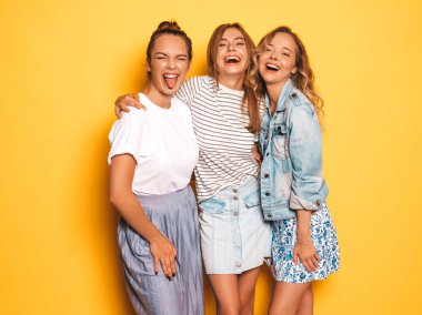 Moda yazlık giysiler üç genç güzel gülümseyen hipster kız. Seksi kaygısız kadınlar stüdyoda sarı duvar yakınında poz. Pozitif modeller deli gidiyor ve eğleniyor. Dili gösterme