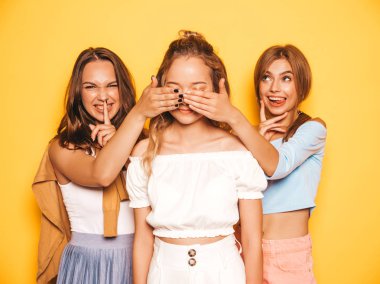 Moda yazlık giysiler üç genç güzel gülümseyen hipster kız. Seksi kaygısız kadınlar stüdyoda sarı duvar yakınında poz. Pozitif modeller onun kadın en iyi arkadaşı şaşırtıcı. Gözlerini kapıyor lar ve arkadan sarılırlar.
