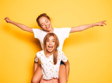 Trendy yaz giysileri iki genç güzel gülümseyen hipster kız. Seksi kaygısız kadınlar stüdyoda sarı duvar yakınında poz. Model arkadaşının sırtında oturan ve ellerini kaldırarak