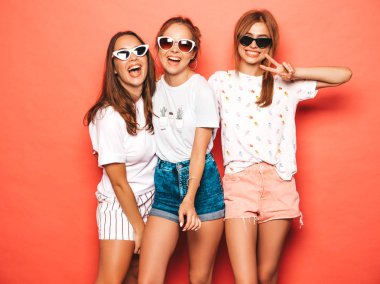 Moda yazlık giysiler üç genç güzel gülümseyen hipster kız. Seksi kaygısız kadınlar stüdyoda pembe duvar yakınında poz. Pozitif modeller deli gidiyor ve güneş gözlüğü eğlenmek
