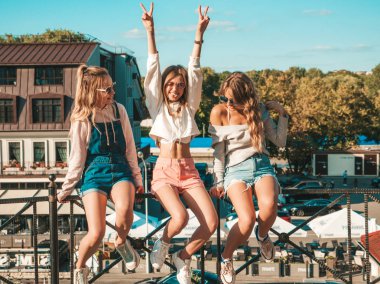 Moda yazlık giysiler üç genç güzel gülümseyen hipster kız Portresi. Sokakta ki tırabzanda oturan seksi kaygısız kadınlar. Güneş gözlüğü ile eğlenen pozitif modeller. El kaldırıyorlar.