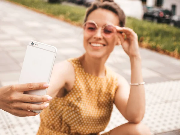 夏季时髦黄色连衣裙中美丽的微笑黑发女孩的特写肖像 模型在智能手机上自拍 女人在温暖的阳光明媚的日子在街上用太阳镜拍照 — 图库照片