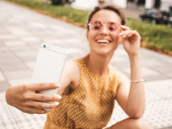 夏季时髦黄色连衣裙中美丽的微笑黑发女孩的特写肖像 模型在智能手机上自拍 女人在温暖的阳光明媚的日子在街上用太阳镜拍照 — 图库照片