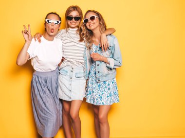 Moda yazlık giysiler üç genç güzel gülümseyen hipster kız. Seksi kaygısız kadınlar stüdyoda sarı duvar yakınında poz. Pozitif modeller deli gidiyor ve güneş gözlüğü eğleniyor. Dillerini gösteriyorlar.