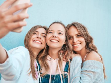 Yazlık kıyafetler giyen üç genç gülümseyen hipster kadın. Akıllı telefonda selfie selfie portre fotoğrafları çeken kızlar. Duvara yakın sokakta poz veren mankenler. Kadın olumlu yüz duyguları gösteren