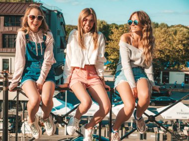 Moda yazlık giysiler üç genç güzel gülümseyen hipster kız. Sokakta parmaklıklarüzerinde oturan seksi kadınlar. Güneş gözlüğü ile eğlenen pozitif modeller. İletişim kuruyorlar ve bir şeyi tartışıyorlar.