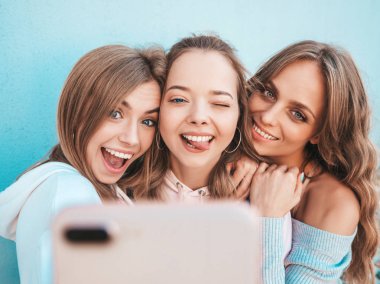 Yazlık kıyafetler giyen üç genç gülümseyen hipster kadın. Akıllı telefonda selfie selfie portre fotoğrafları çeken kızlar. Duvara yakın sokakta poz veren mankenler. Kadın olumlu yüz duyguları gösteren