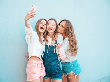 Yazlık kıyafetler giyen üç genç gülümseyen hipster kadın. Akıllı telefonda selfie selfie portre fotoğrafları çeken kızlar. Duvara yakın sokakta poz veren mankenler. Kadın olumlu yüz duyguları gösteriyor. Dili gösterme
