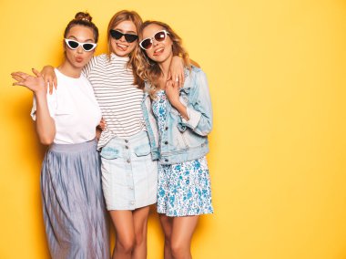 Moda yazlık giysiler üç genç güzel gülümseyen hipster kız. Seksi kaygısız kadınlar stüdyoda sarı duvar yakınında poz. Pozitif modeller deli gidiyor ve güneş gözlüğü eğlenmek