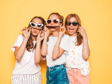 Moda yazlık giysiler üç genç güzel gülümseyen hipster kız. Seksi kaygısız kadınlar stüdyoda sarı duvar yakınında poz. Pozitif modeller deli gidiyor ve eğleniyor. Saç ile bıyık yapma