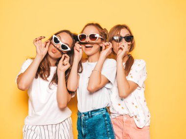 Moda yazlık giysiler üç genç güzel gülümseyen hipster kız. Seksi kaygısız kadınlar stüdyoda sarı duvar yakınında poz. Pozitif modeller deli gidiyor ve eğleniyor. Saç ile bıyık yapma