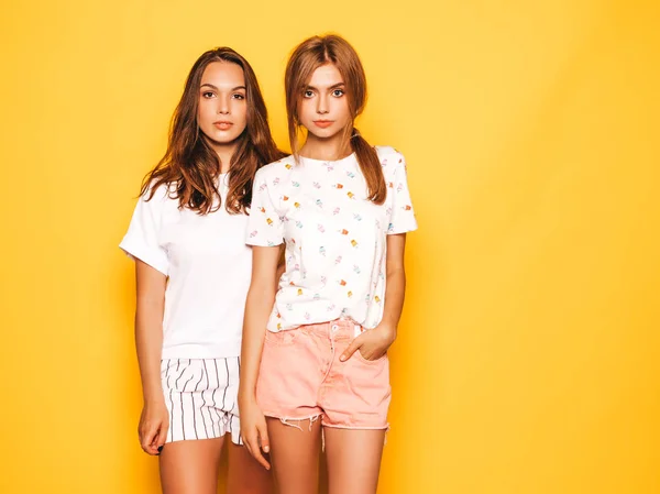 Two young beautiful boring hipster girls in trendy summer clothes. Sexy carefree women posing near yellow wall in studio