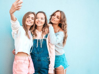 Yazlık kıyafetler giyen üç genç gülümseyen hipster kadın. Akıllı telefonda selfie selfie portre fotoğrafları çeken kızlar. Duvara yakın sokakta poz veren mankenler. Kadın olumlu yüz duyguları gösteren.