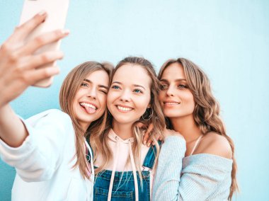 Yazlık kıyafetler giyen üç genç gülümseyen hipster kadın. Akıllı telefonda selfie selfie portre fotoğrafları çeken kızlar. Duvara yakın sokakta poz veren mankenler. Kadın olumlu yüz duyguları gösteren.
