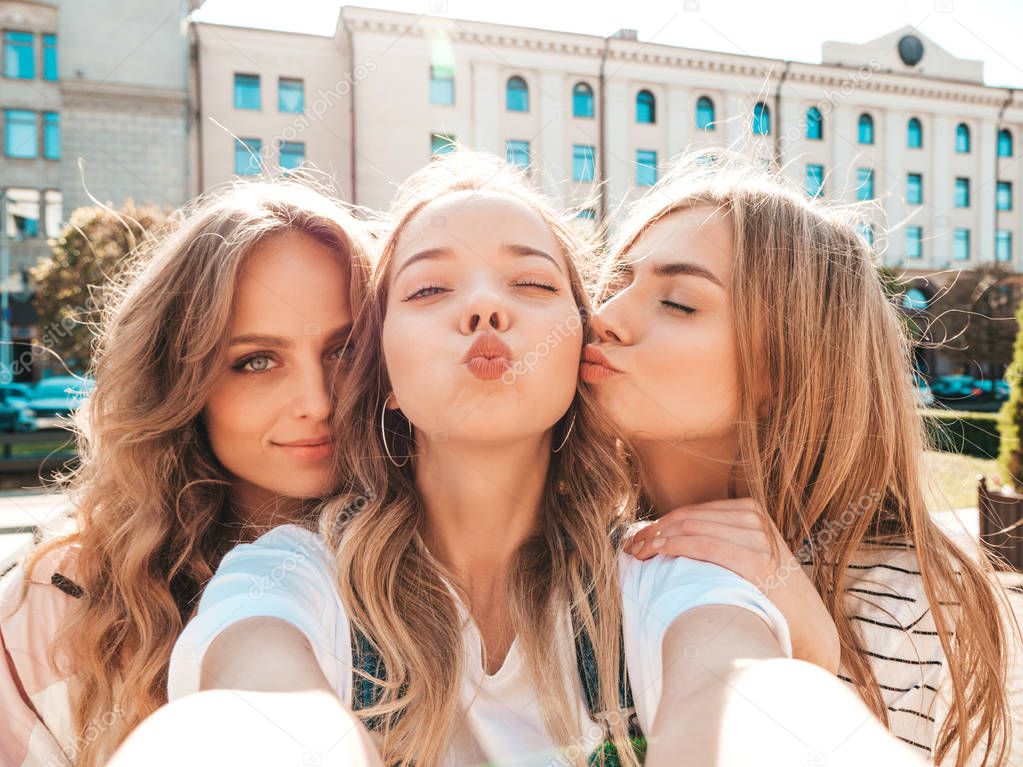 Three young smiling hipster women in summer clothes.Girls taking selfie self portrait photos on smartphone.Models posing in the street.Female kissing their friend in cheek