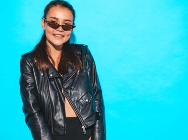 Moda yamsız siyah deri ceket ve güneş gözlüğü genç güzel hipster kötü kız Portresi. Seksi kaygısız kadın mavi izole. Esmer model stüdyoda poz