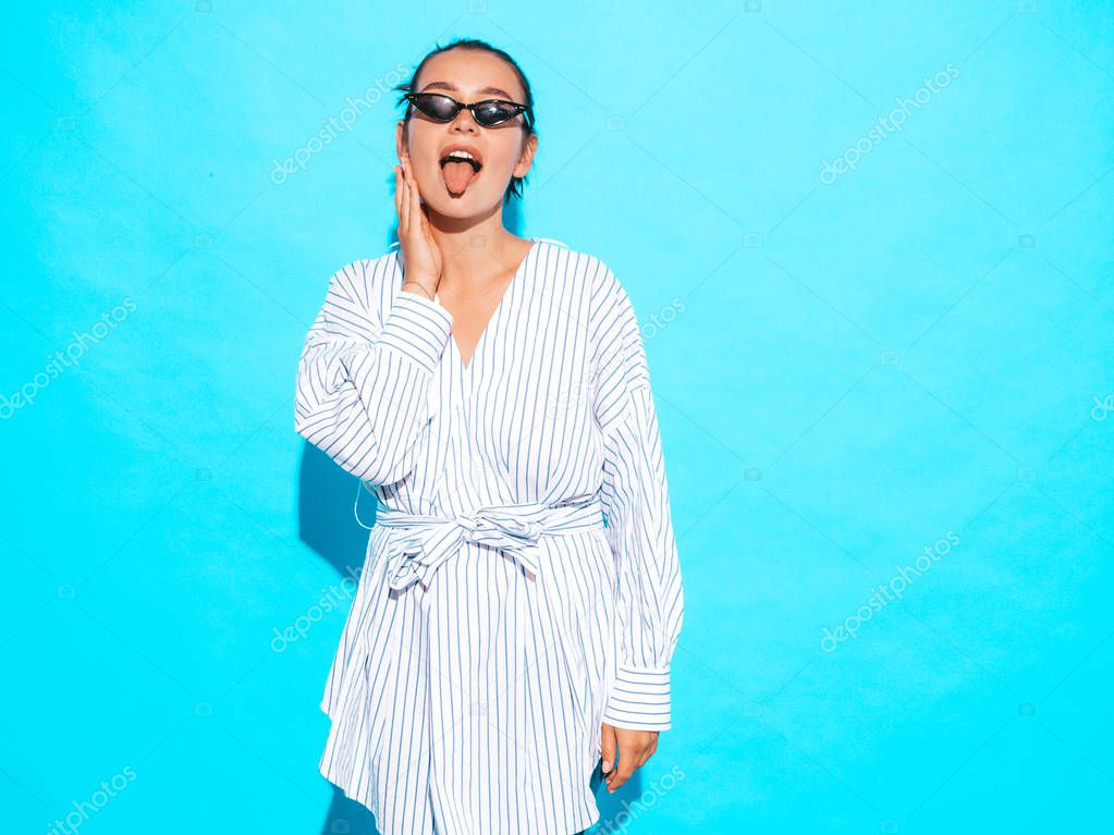 Portrait of young beautiful smiling hipster girl in trendy summer clothes. Sexy carefree woman posing near blue wall. Positive model having fun in sunglasses.Shows tongue