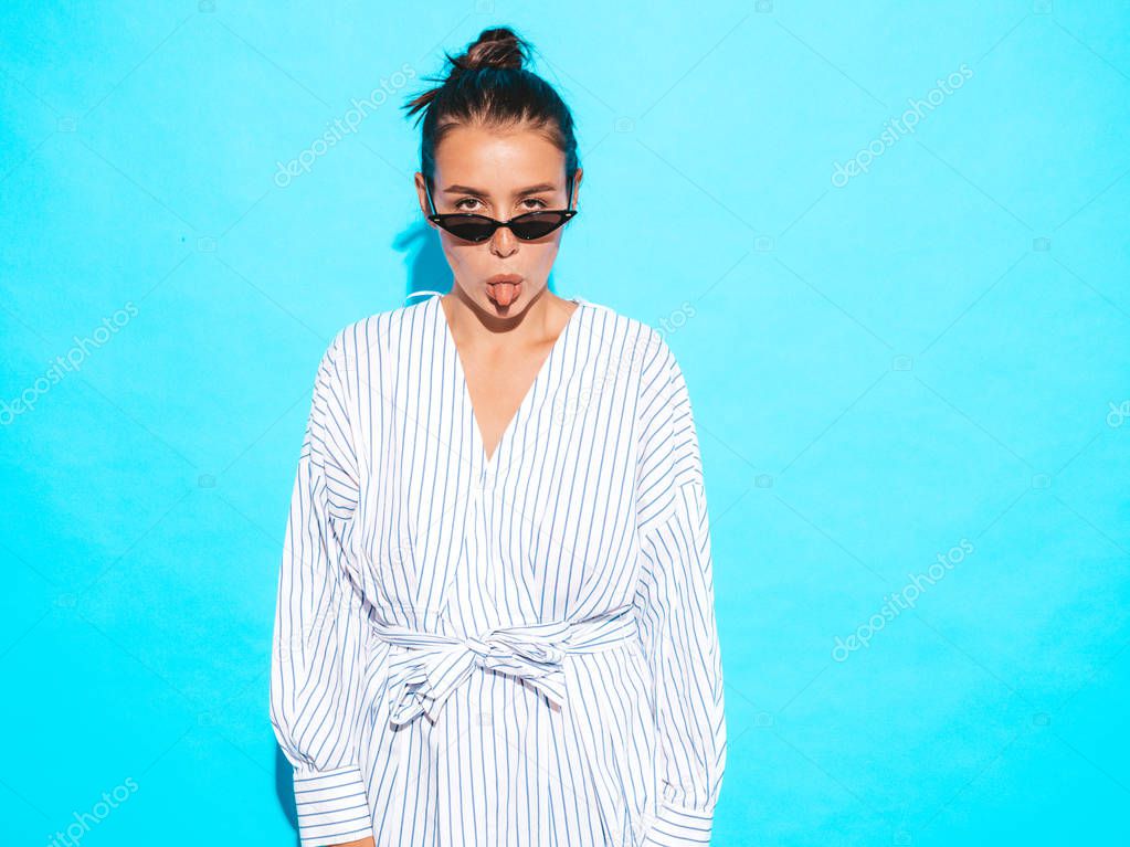 Portrait of young beautiful smiling hipster girl in trendy summer clothes. Sexy carefree woman posing near blue wall. Positive model having fun in sunglasses.Shows tongue