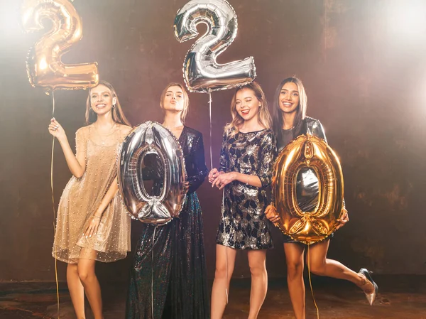 Beautiful Women Celebrating New Year. Happy Gorgeous Girls In Stylish Sexy Party Dresses Holding Gold and Silver 2020 Balloons, Having Fun At New Year\'s Eve Party. Holiday Celebration.Charming Models