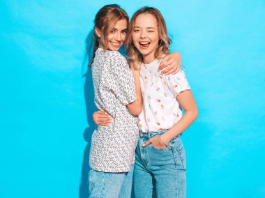 İki güzel, gülümseyen sarışın hippi kız yazın moda renkli tişört giyiyorlar. Mavi duvarın yanında poz veren kaygısız seksi kadınlar. Pozitif modeller eğleniyor ve dil gösteriyor.