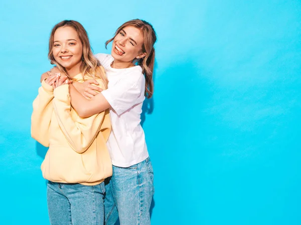 两个年轻美丽的金发嬉皮士女孩穿着时髦的夏天艳丽的T恤衫 在蓝色墙边摆姿势的无忧无虑的女人 有乐趣的正面模型和显示舌头的正面模型 — 图库照片