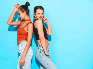 İki güzel, gülümseyen esmer hippi kız aynı moda yaz kıyafetleri içinde. Stüdyoda mavi duvarın yanında poz veren kaygısız kadınlar. Pozitif mankenler eğleniyor.