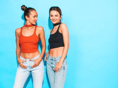 İki güzel, gülümseyen esmer hippi kız aynı moda yaz kıyafetleri içinde. Stüdyoda mavi duvarın yanında poz veren kaygısız kadınlar. Pozitif mankenler eğleniyor.