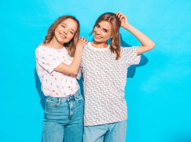 İki genç trendy sarışın hipster kızlarda gülümseyen güzel renkli tişört elbise yaz. Seksi kaygısız kadın mavi duvara yakın poz. Olumlu modelleri eğleniyor