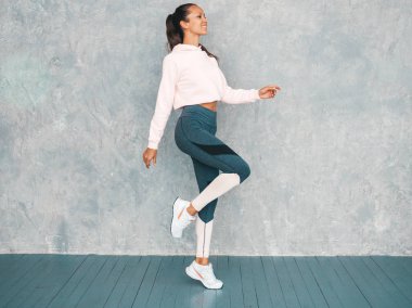 Spor kıyafetleri içindeki kendine güvenen kadın portresi. Spor giyimli genç bir bayan. Mükemmel bronzlaşmış vücutlu güzel bir model. Stüdyoda gri duvarın yanında koşan bir kadın.