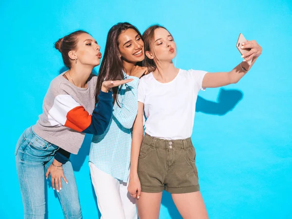 夏の服で3人の若い笑顔ヒップスター女性 スマートフォンで自撮り自画像写真を撮る女の子 スタジオで青い壁の近くにポーズモデル 肯定的な感情を示す女性 — ストック写真
