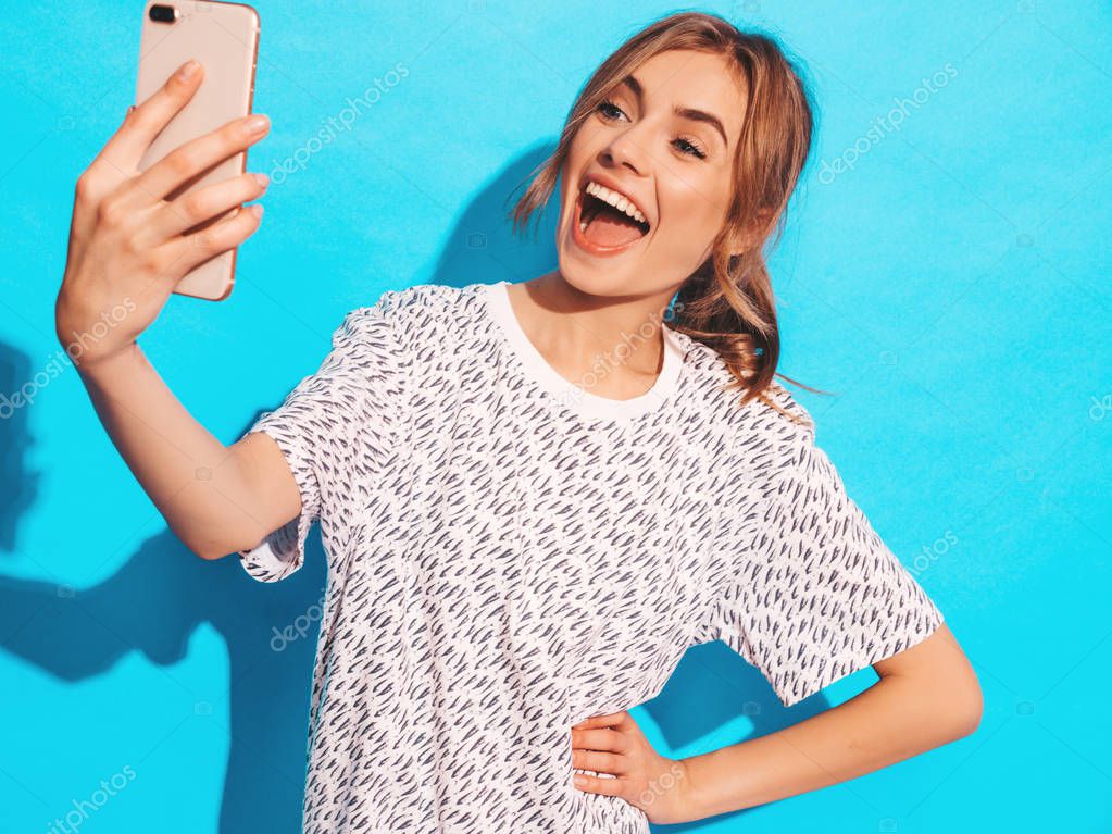 Portrait of cheerful young woman taking photo selfie. Beautiful girl holding smartphone camera. Smiling model posing near blue wall in studio. Surprised model shocked