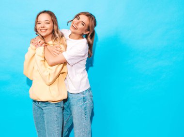 İki genç trendy sarışın hipster kızlarda gülümseyen güzel renkli tişört elbise yaz. Seksi kaygısız kadın mavi duvara yakın poz. Olumlu modelleri eğleniyor 