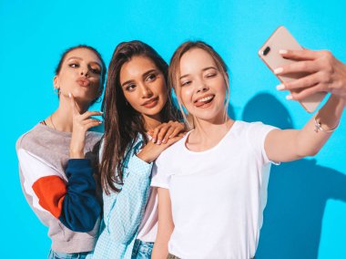 Three young smiling hipster women in summer clothes.Girls taking selfie self portrait photos on smartphone.Models posing near blue wall in studio.Female showing positive emotions