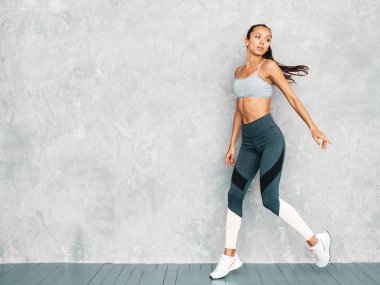 Spor kıyafetleri içindeki kendine güvenen kadın portresi. Spor giyimli genç bir bayan. Mükemmel bronzlaşmış vücutlu güzel bir model. Gri duvarın yanındaki stüdyoda atlayan bir kadın.