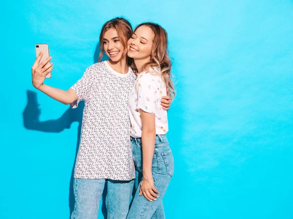 2人の若いスタイリッシュな笑顔ブロンドの女性の肖像画 夏のヒップスター服を着た女の子 スタジオの青い壁の近くのスマートフォンでセルフィーを作る肯定的なモデル — ストック写真