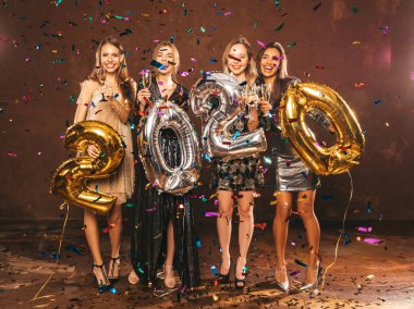 Yeni Yılı Kutlayan Güzel Kadınlar. Altın ve Gümüş Balonlar Tutan Şık Parti Elbiseli Mutlu Güzel Kızlar, Yeni Yıl Partisi 'nde eğleniyor ve şampanya flüt içiyor.