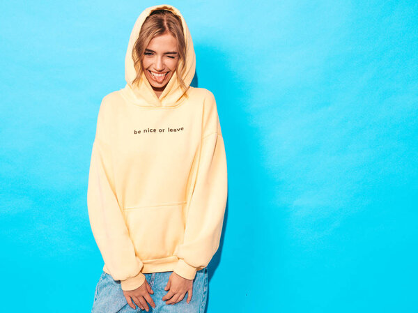 Portrait of young beautiful smiling girl in trendy summer hipster yellow hoodie.Sexy carefree woman posing near blue wall. Positive model having fun