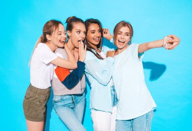 Yaz kıyafetleri içinde dört genç, gülümseyen hippi kadın. Akıllı telefondan selfie çeken kızlar. Stüdyoda mavi duvarın yanında poz veren mankenler.